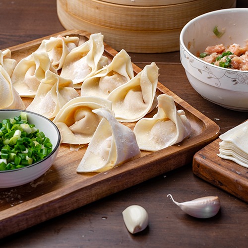 Wooden plate of folded wonton wrappers with bowl of pork meat and spring onions